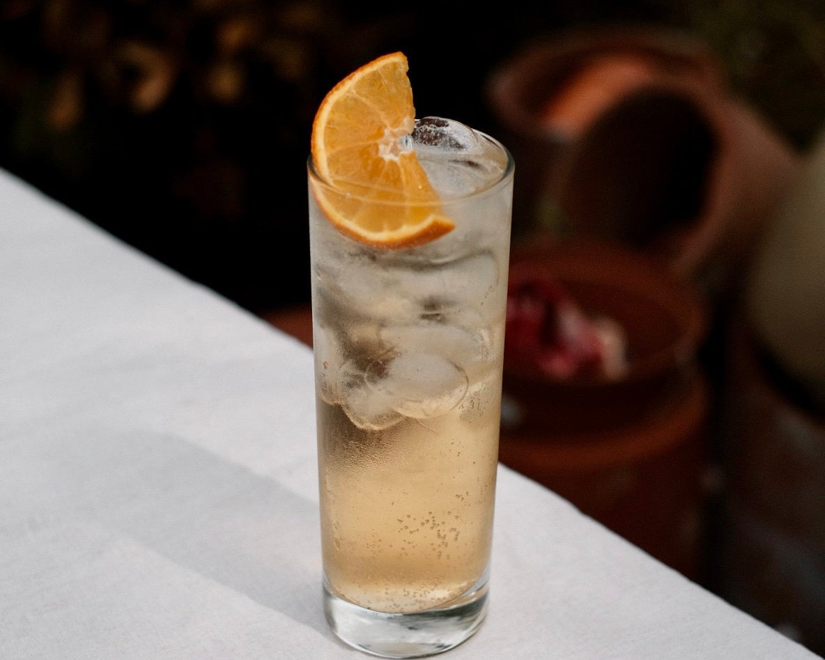 Tall glass of iced golden spiced rum highball garnished with an orange wedge, served outdoors at Loveday Distillery