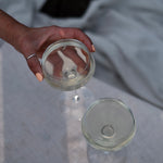 Hand holding a coupe glass of pale Kissing Gate Gin cocktail, second glass on white linen surface, Cornwall