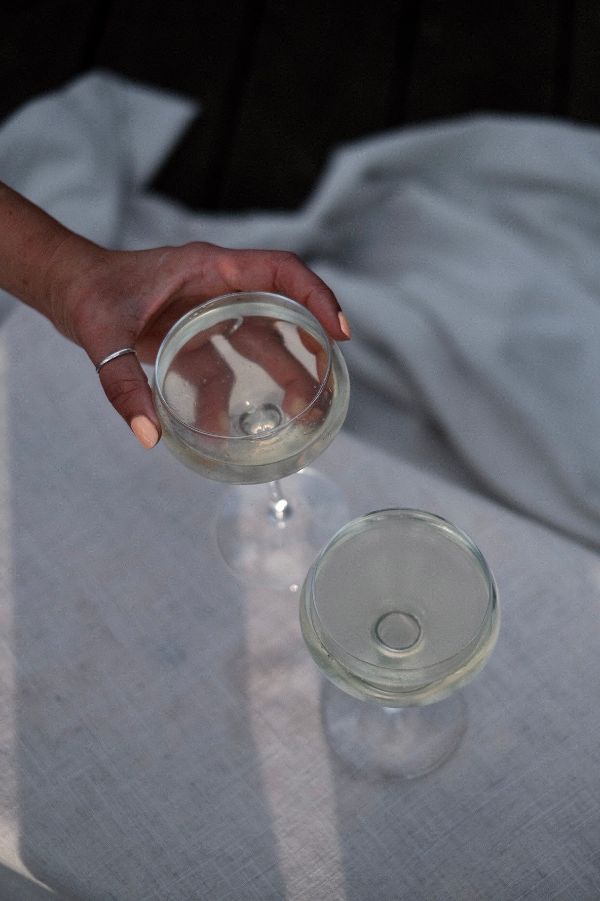 Hand holding a coupe glass of pale Kissing Gate Gin cocktail, second glass on white linen surface, Cornwall