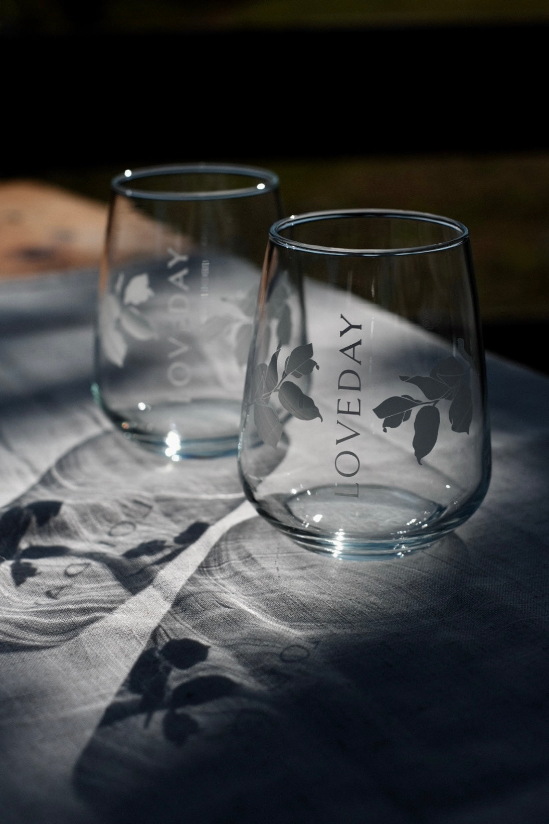 Two etched stemless wine glasses engraved with Loveday branding and leaf motifs, resting on a linen cloth in dappled sunlight
