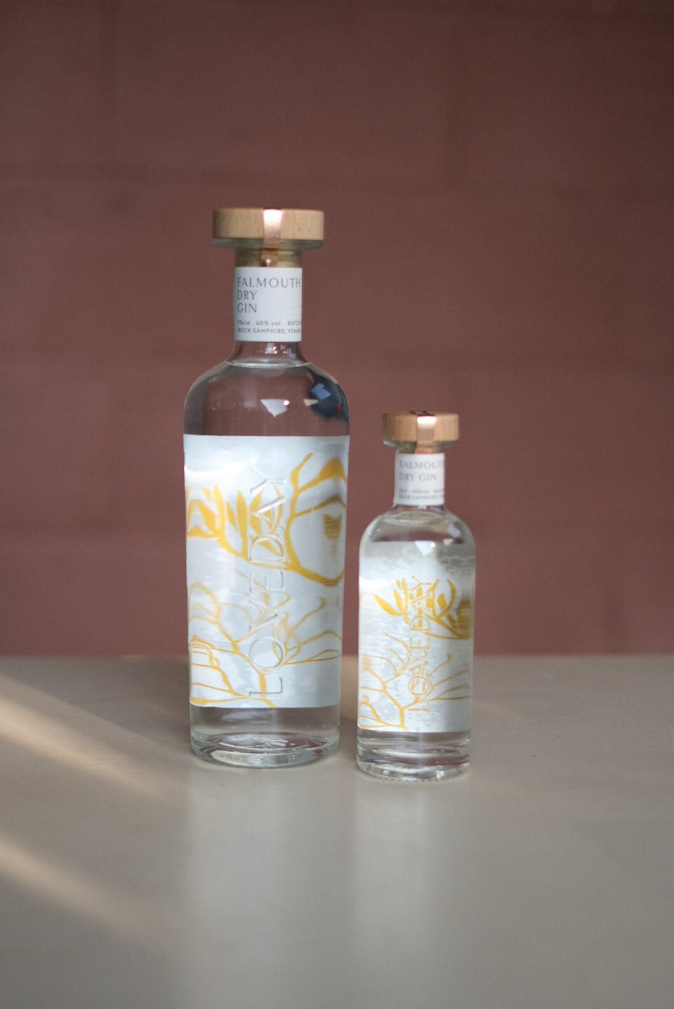 Two Loveday Falmouth Dry Gin bottles, large and mini, clear glass with gold botanical labels and copper caps