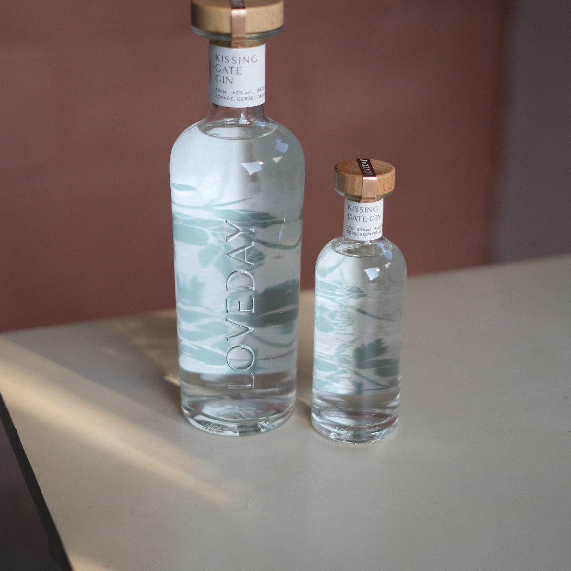Two clear glass Loveday Kissing Gate Gin bottles, large and mini, with wooden caps and teal wave design, on a pale surface
