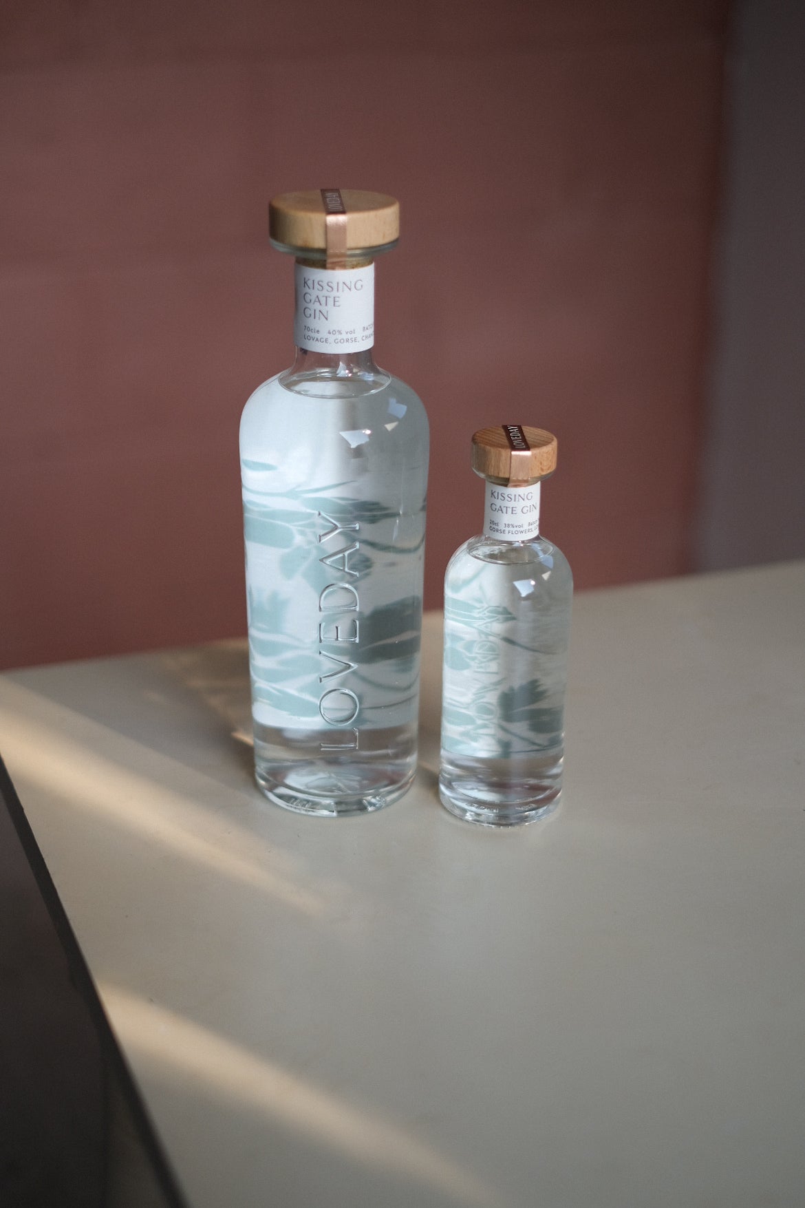 Two clear glass Loveday Kissing Gate Gin bottles, large and mini, with wooden caps and teal wave design, on a pale surface