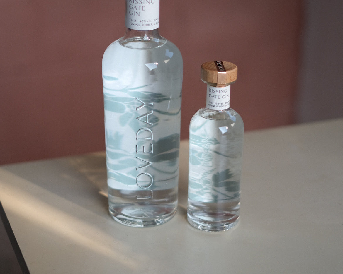 Two clear glass Loveday Kissing Gate Gin bottles, large and mini, with wooden caps and teal wave design, on a pale surface