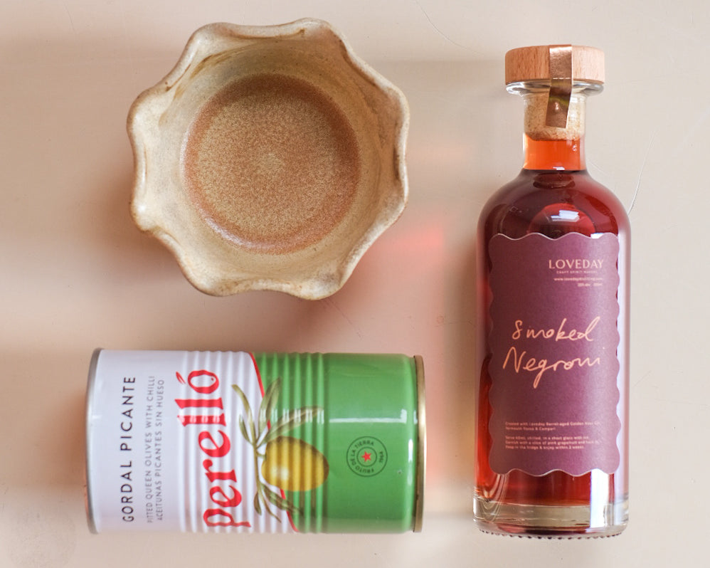 Loveday Smoked Negroni bottle, Perelló Gordal Picante olive tin, and a rustic ceramic bowl on a pale pink surface