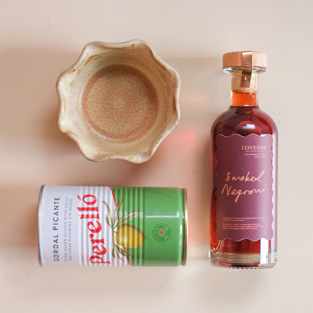 Loveday Smoked Negroni bottle, Perelló Gordal Picante olive tin, and a rustic ceramic bowl on a pale pink surface
