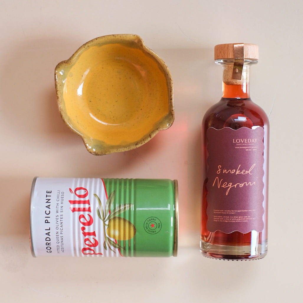 Loveday Smoked Negroni bottle, Perelló Gordal Picante olive tin, and yellow ceramic bowl on pale pink surface