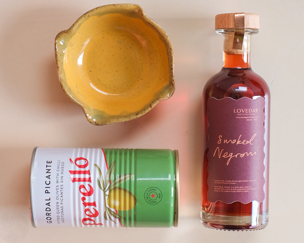 Loveday Smoked Negroni bottle, Perelló Gordal Picante olive tin, and yellow ceramic bowl on pale pink surface