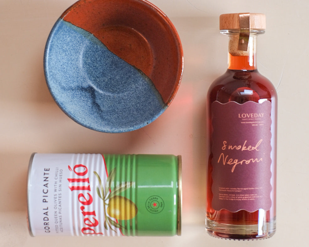 Loveday Smoked Negroni bottle with Perelló Gordal Picante olive tin and blue-terracotta ceramic bowl on beige surface