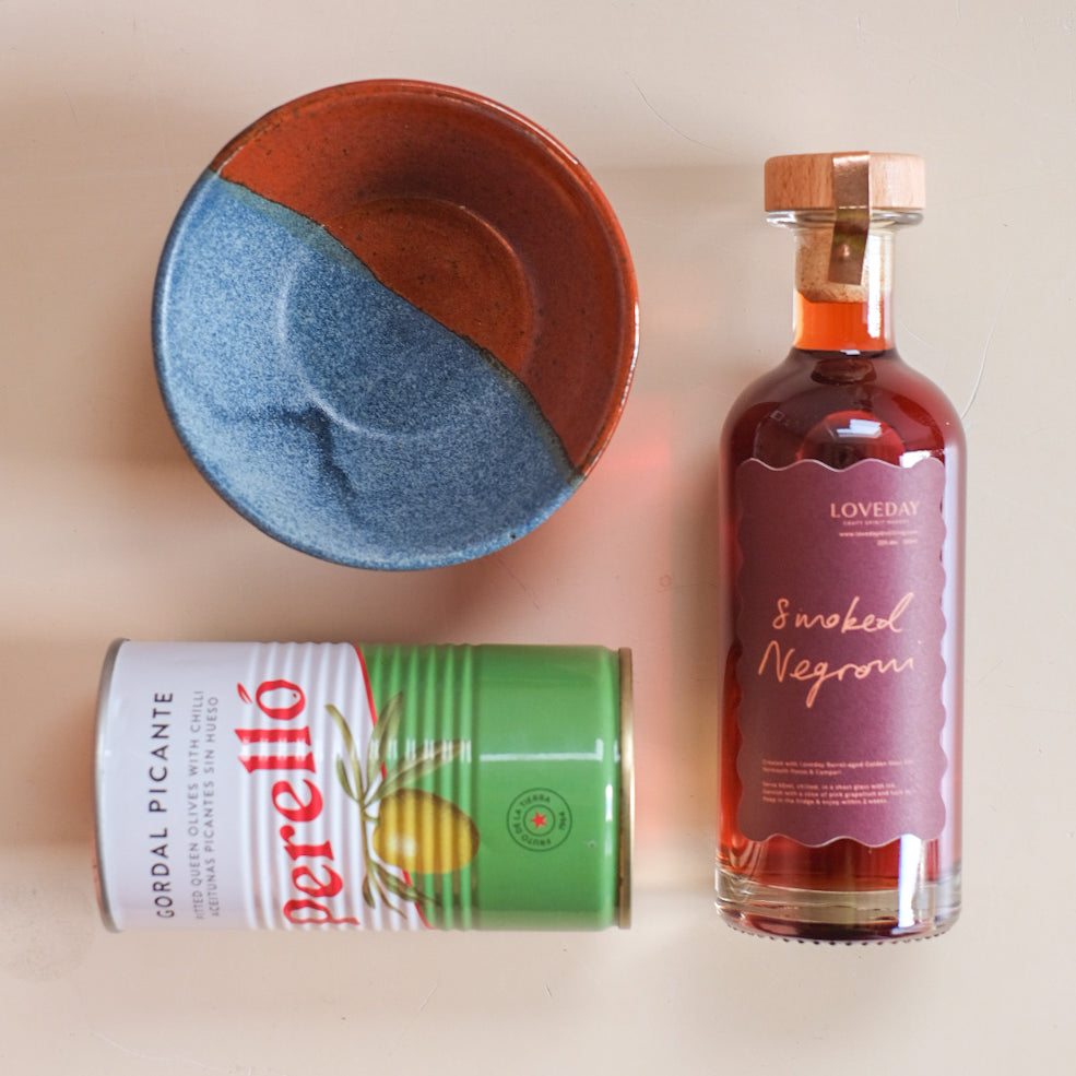 Loveday Smoked Negroni bottle with Perelló Gordal Picante olive tin and blue-terracotta ceramic bowl on beige surface