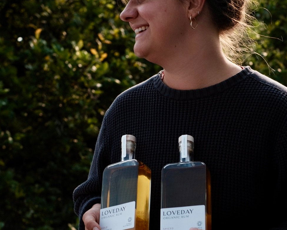 Woman smiling outdoors holding two bottles of Loveday Organic Rum — Gold and Spiced — in a garden setting