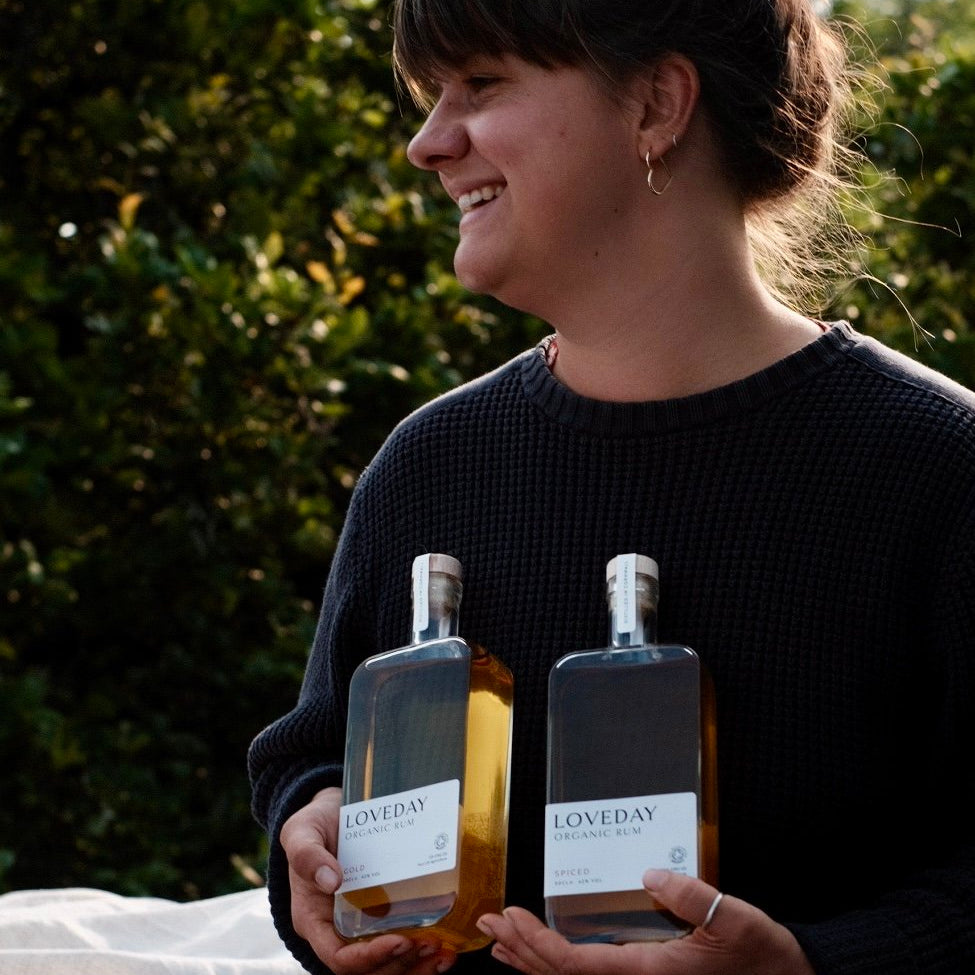 Woman smiling outdoors holding two bottles of Loveday Organic Rum — Gold and Spiced — in a garden setting