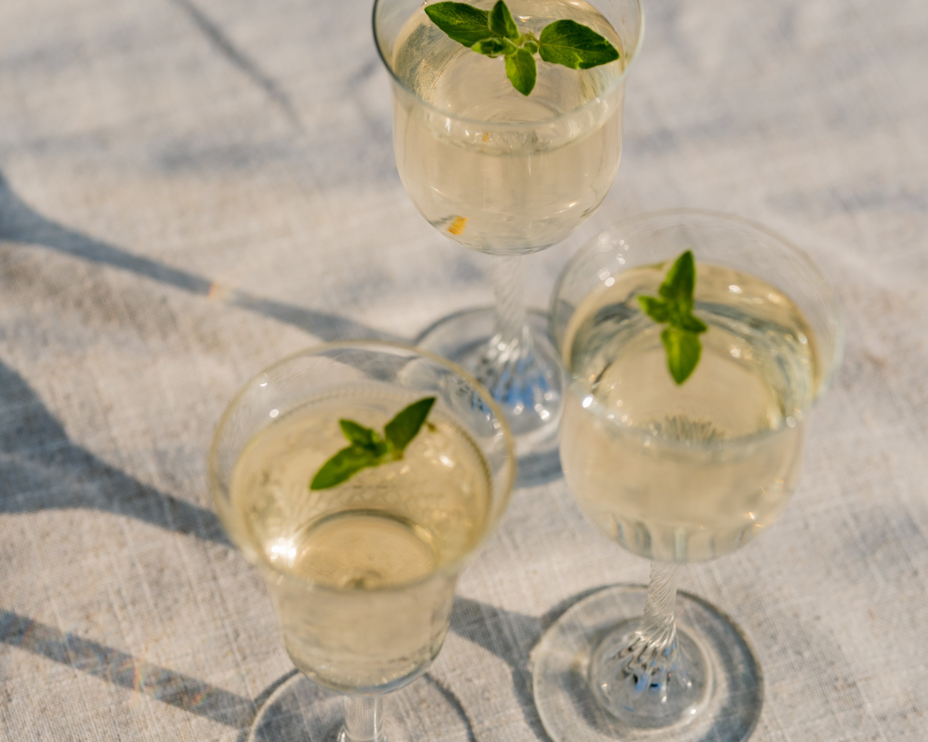 Three stemmed glasses of pale gold Loveday Distillery gin garnished with fresh herb sprigs on a linen cloth in sunlight