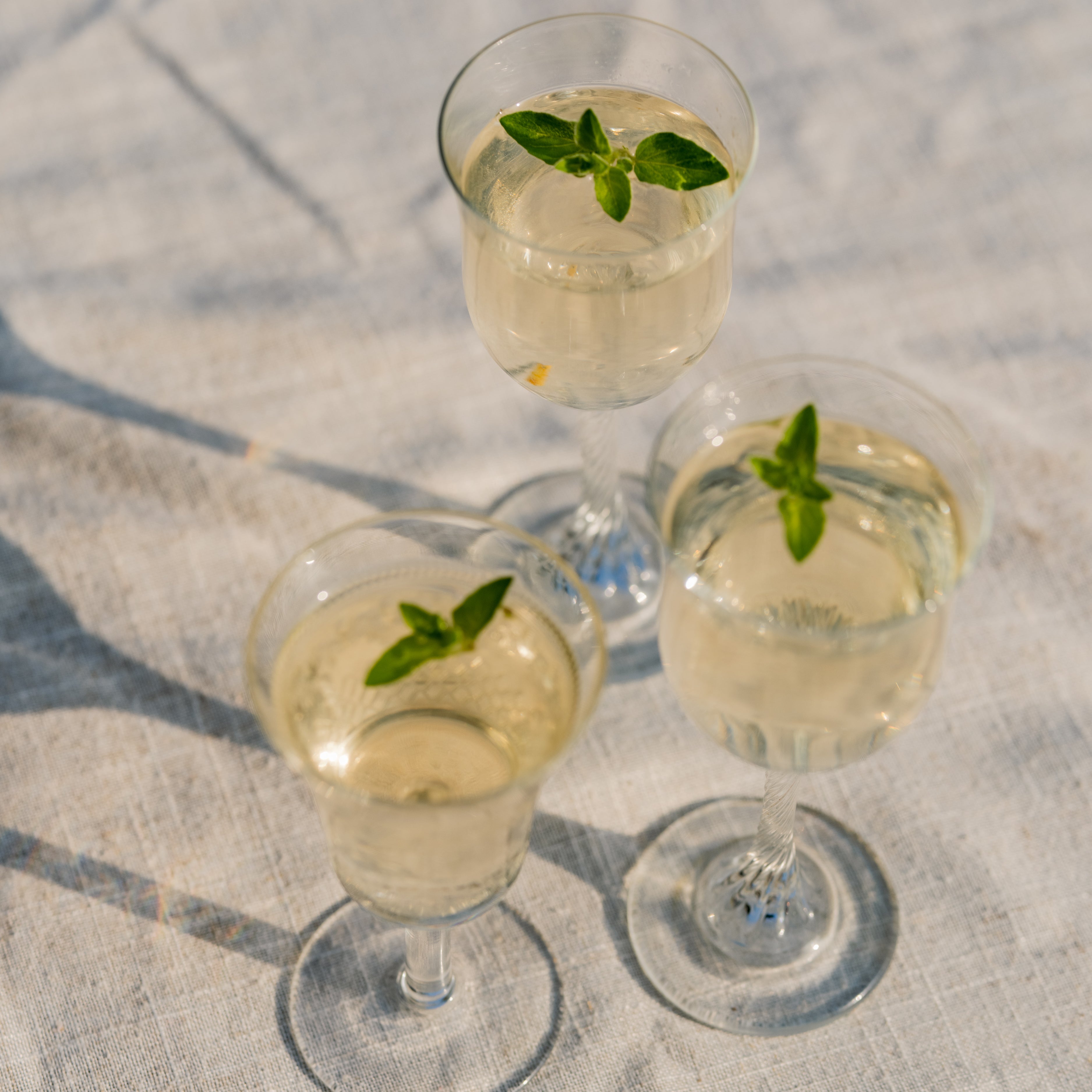 Three stemmed glasses of pale gold Loveday Distillery gin garnished with fresh herb sprigs on a linen cloth in sunlight