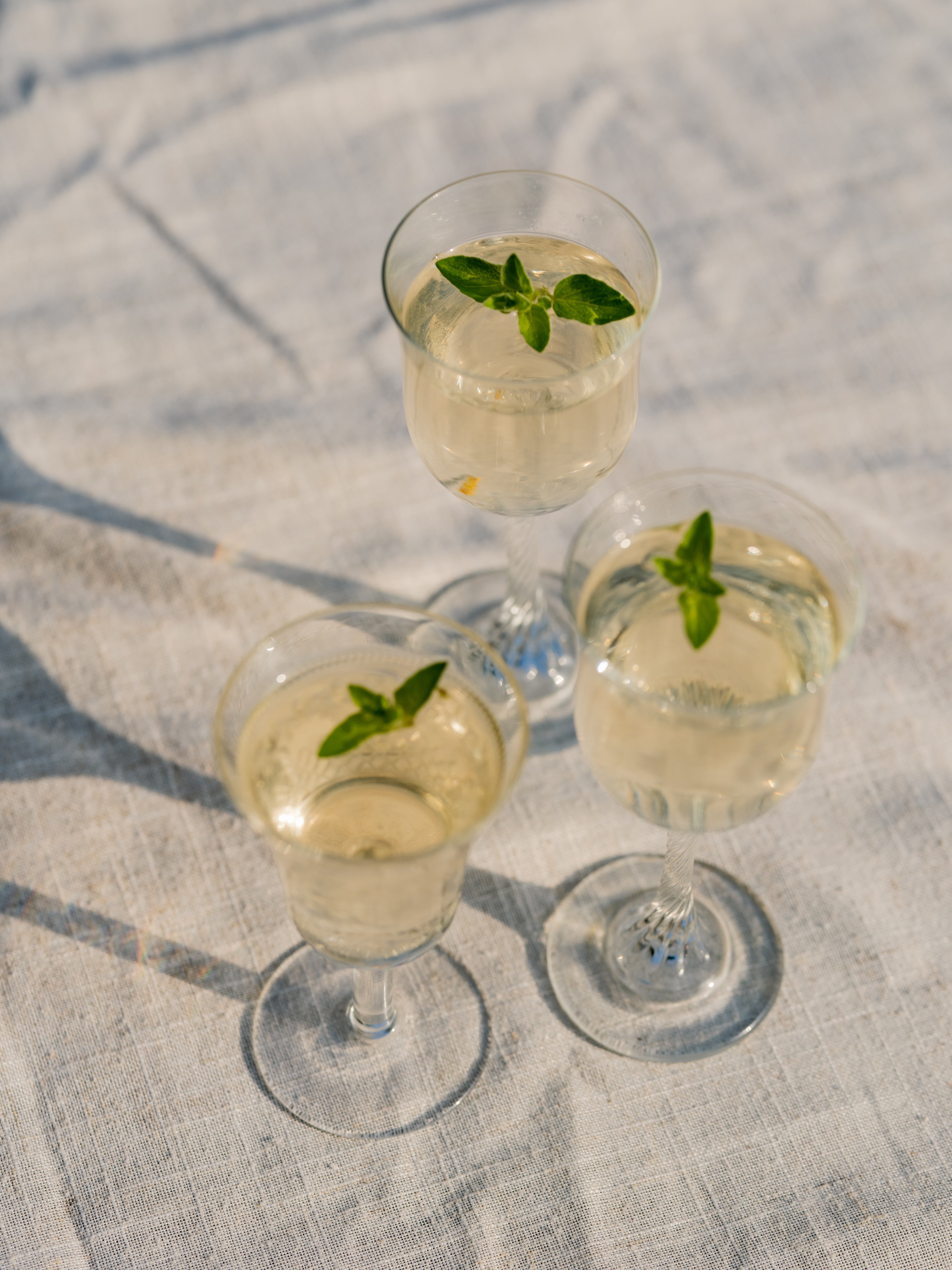 Three stemmed glasses of pale gold Loveday Distillery gin garnished with fresh herb sprigs on a linen cloth in sunlight