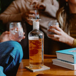 Loveday Golden Hour Gin bottle on wooden table, friends sharing grapefruit gin in stemless glasses at a cosy gathering