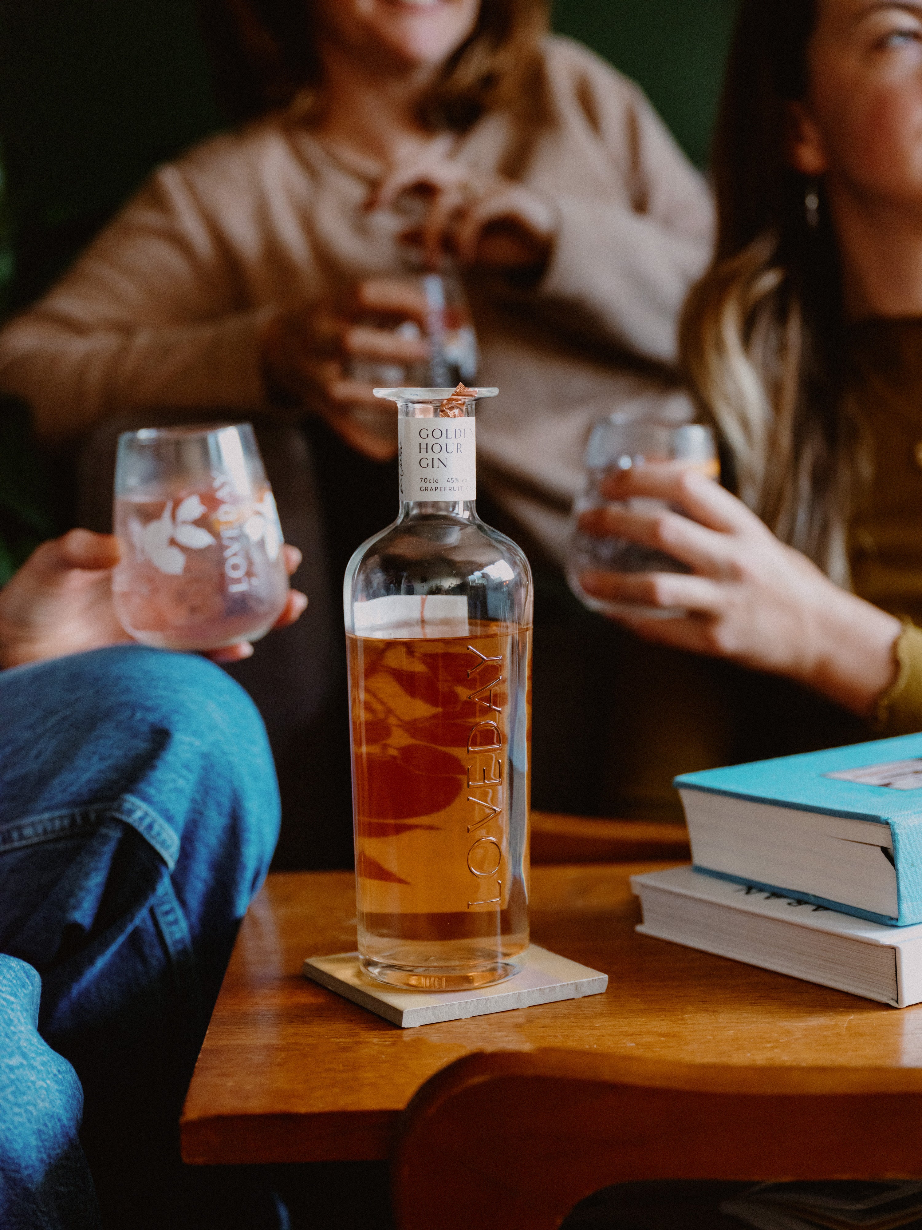 Loveday Golden Hour Gin bottle on wooden table, friends sharing grapefruit gin in stemless glasses at a cosy gathering