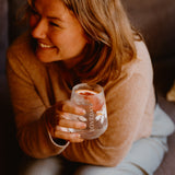 Woman smiling, holding a Loveday stemless glass with ice and a citrus slice in warm golden light