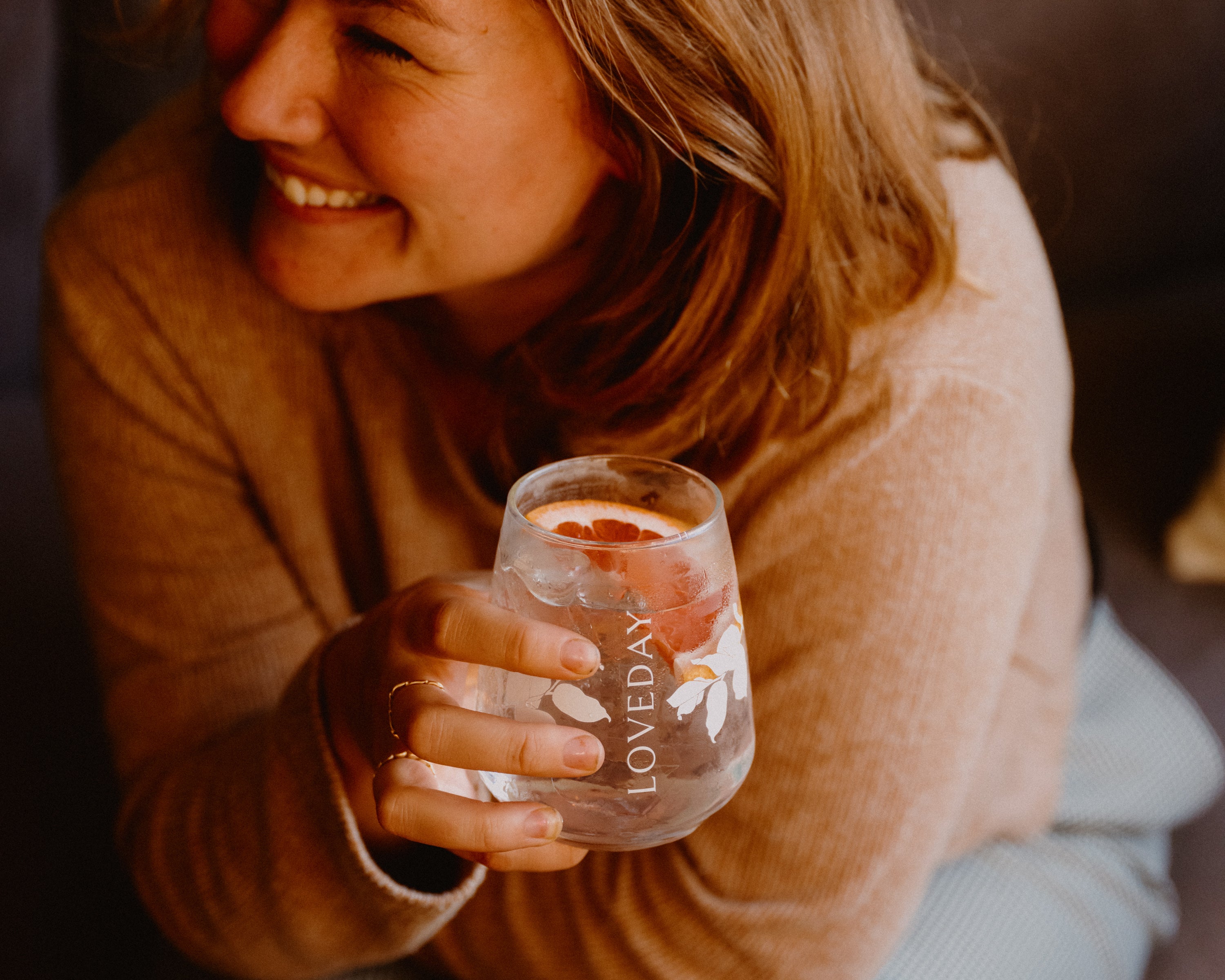 Woman smiling, holding a Loveday stemless glass with ice and a citrus slice in warm golden light