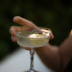 Hand holding a chilled coupe glass of pale golden gin cocktail with herb garnish, outdoors in soft evening light