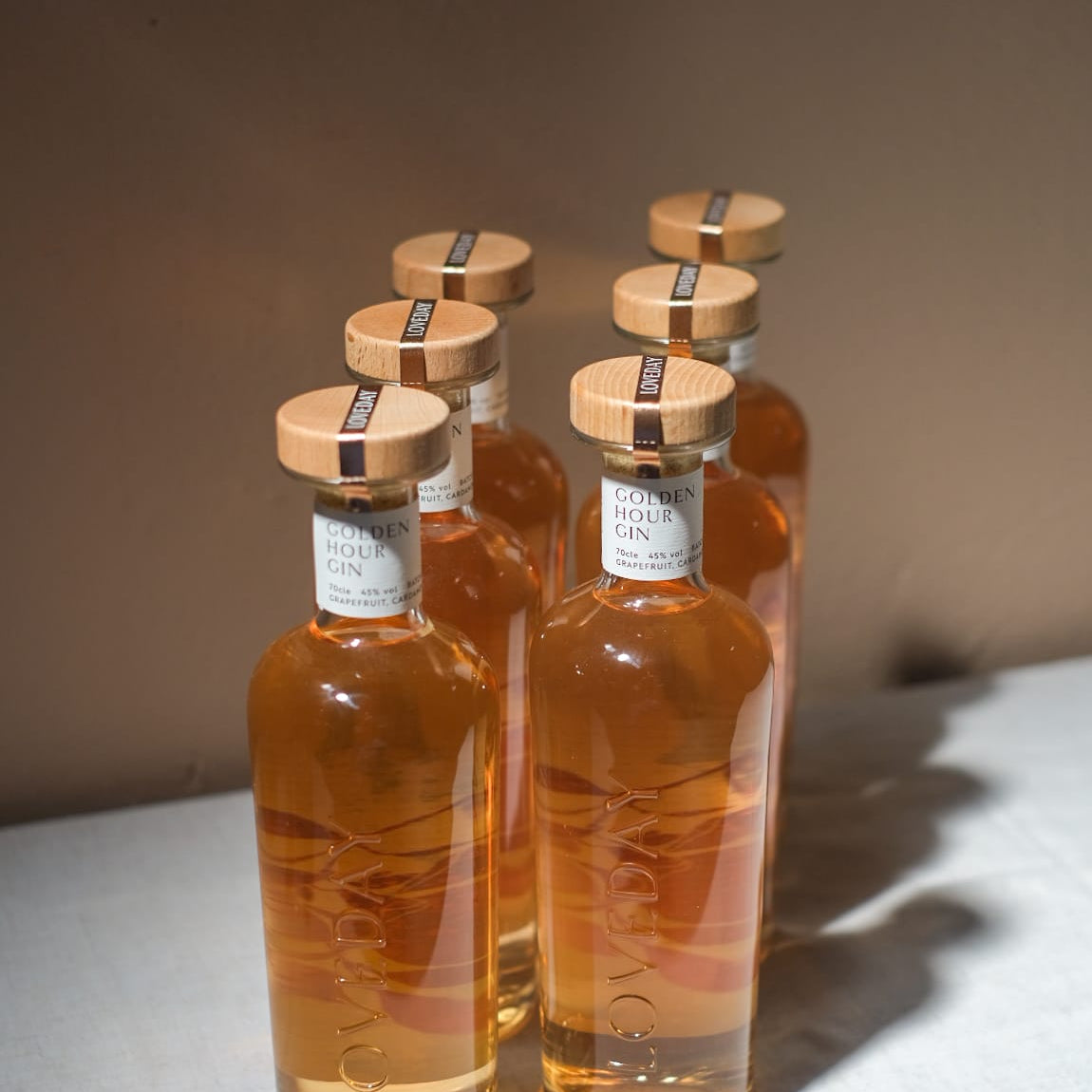 Five bottles of Loveday Golden Hour Gin with wooden caps, amber liquid, arranged in a group on a white surface
