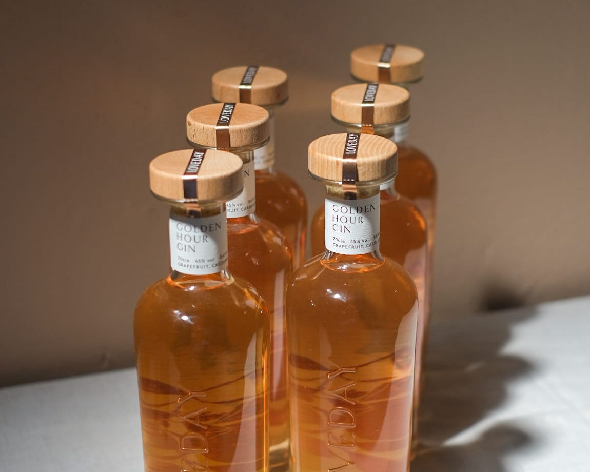 Five bottles of Loveday Golden Hour Gin with wooden caps, amber liquid, arranged in a group on a white surface
