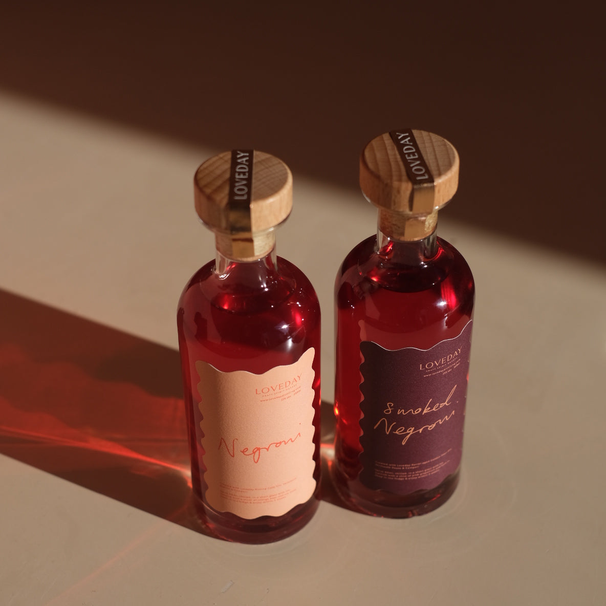 Two Loveday bottled Negroni and Smoked Negroni bottles with wooden stoppers, glowing ruby red in warm directional light