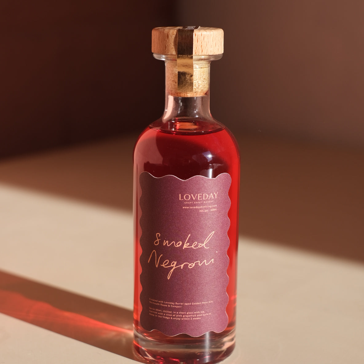 Loveday Smoked Negroni bottle with cork stopper, deep red liquid, scallop-edged mauve label, warm shadow on cream surface