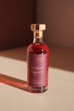 Loveday Smoked Negroni bottle with cork stopper, deep red liquid, scallop-edged mauve label, warm shadow on cream surface