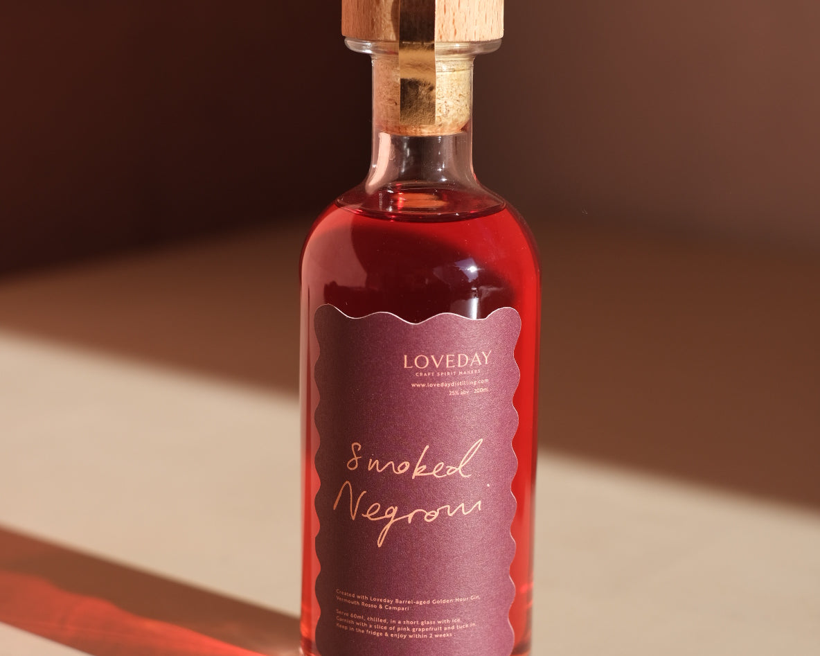 Loveday Smoked Negroni bottle with cork stopper, deep red liquid, scallop-edged mauve label, warm shadow on cream surface